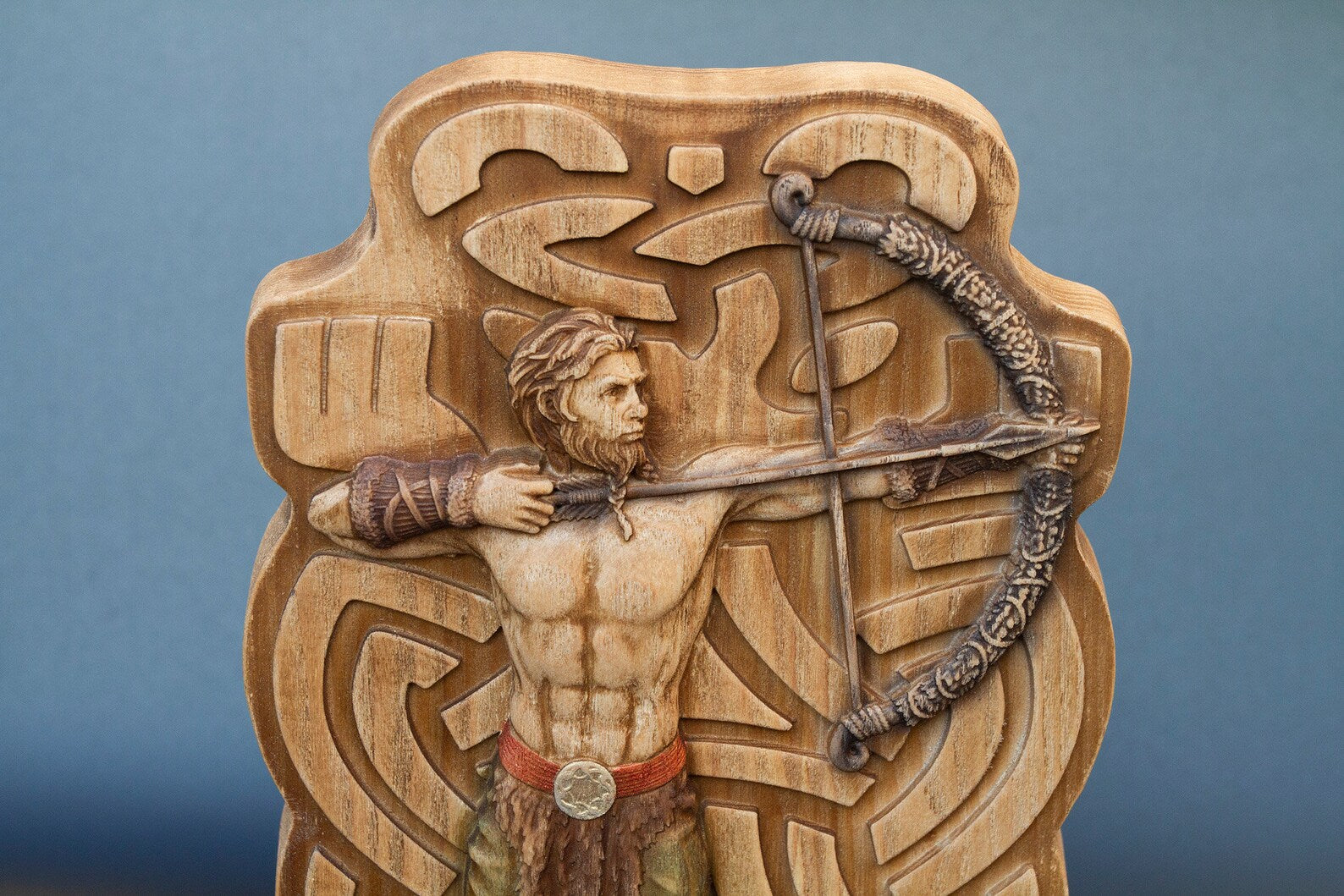 Closeup of wooden Ullr figurine top half showing muscular torso, braided hair, and carved bow against Celtic runic background.